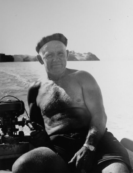 An elderly man with no sits casually in a small boat with an outboard motor.