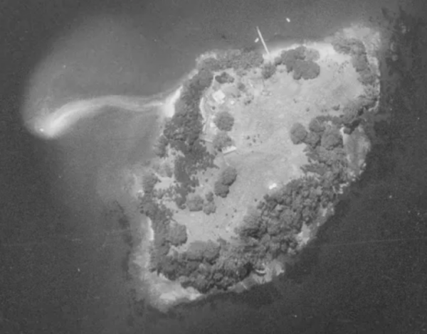 A black and white satellite view of a small island with a large expanse of grass surrounded by vegetation, showing a group of buildings in the top left.