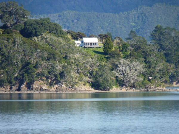 A zoomed view of a house sitting on a hill surrounded by trees with a rocky coastline below and ocean in the foreground.